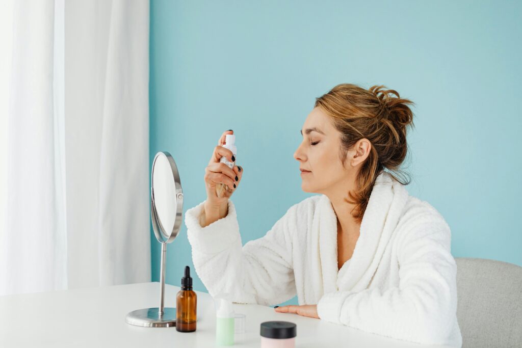 Woman in a bathrobe using a skincare spray in front of a mirror indoors.