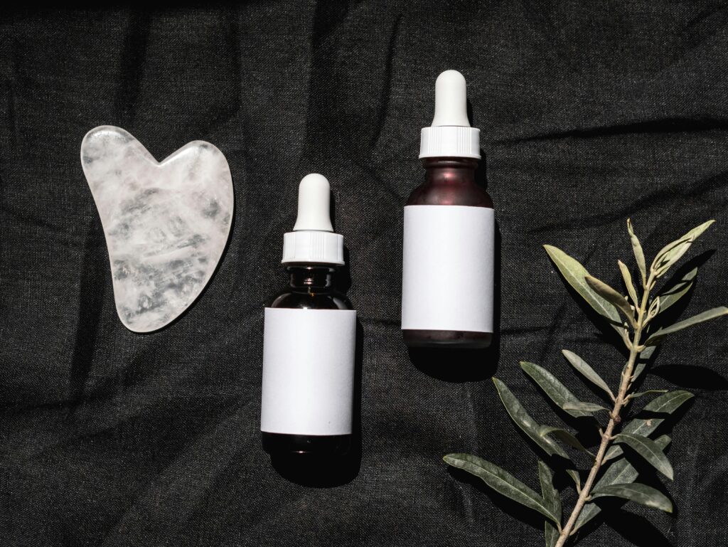 Minimalist flatlay of skincare bottles, gua sha, and a branch on dark fabric background.