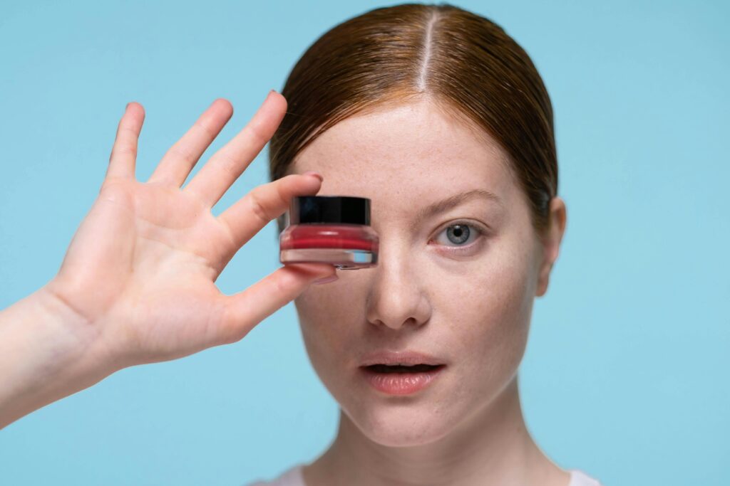 Woman holding a lip balm jar in front of her eye, blue background.